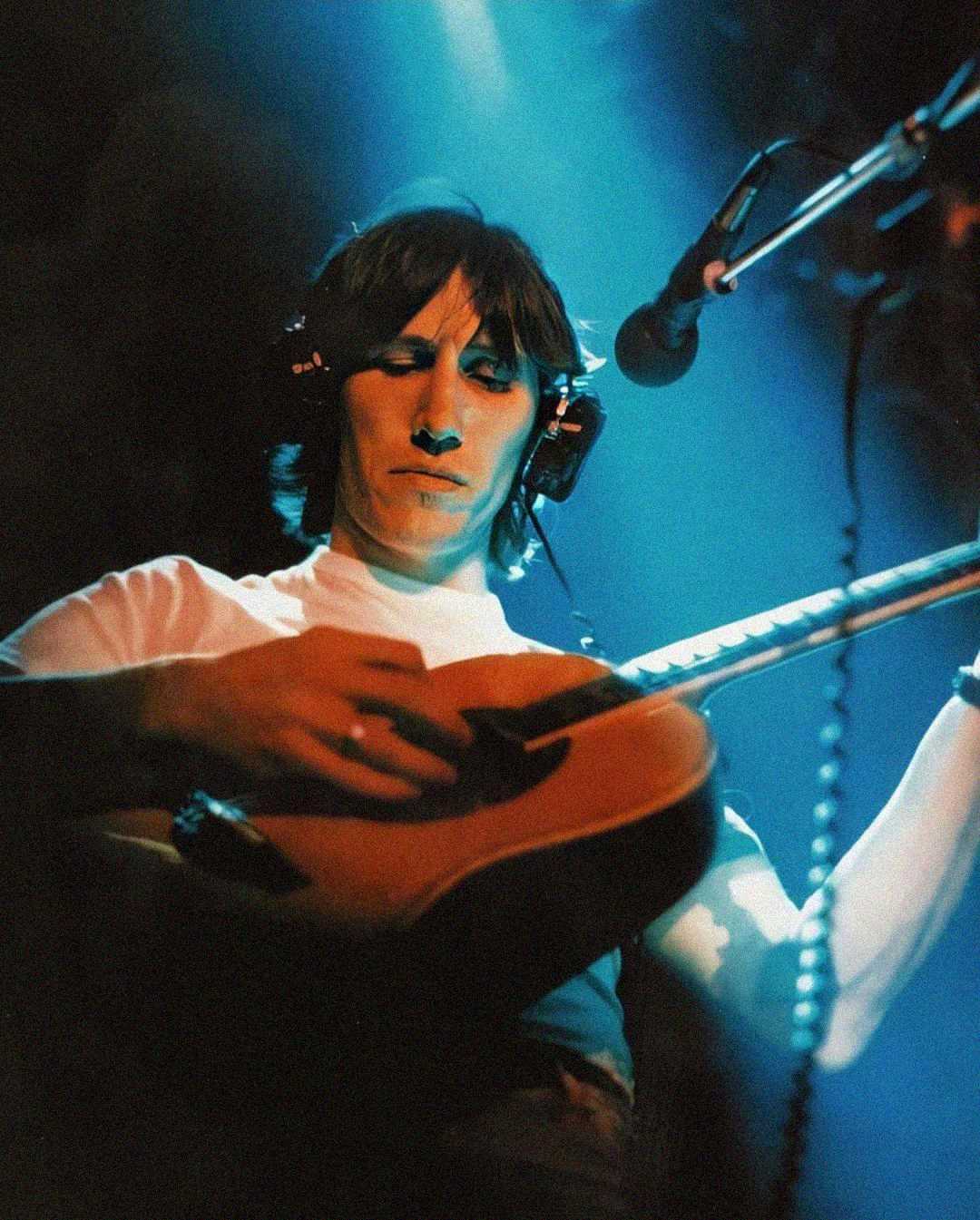 A musician wearing headphones and a white shirt plays an acoustic guitar on stage under dramatic blue and teal lighting, with a microphone positioned nearby.