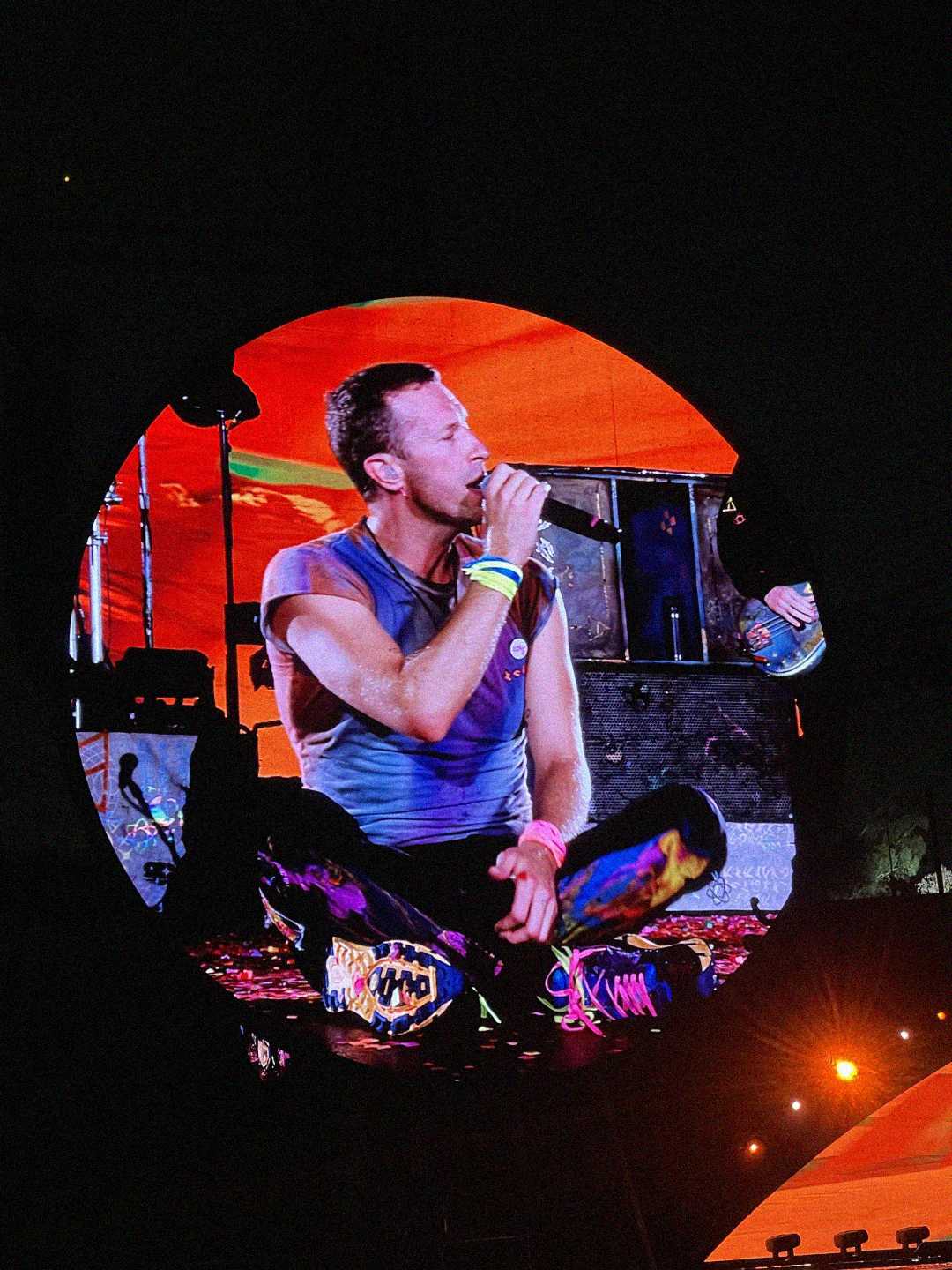 A singer sits cross-legged on stage, holding a microphone and performing in front of a large circular screen with vibrant orange and red colors. He wears a sleeveless shirt and colorful pants. Bright stage lights illuminate the scene.