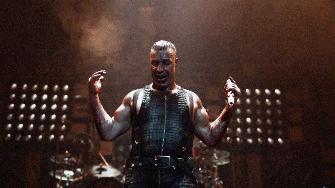 A muscular performer wearing a black leather outfit stands on stage with arms raised, holding a microphone. Dramatic lighting and smoke create an intense atmosphere, with drum equipment visible in the background.