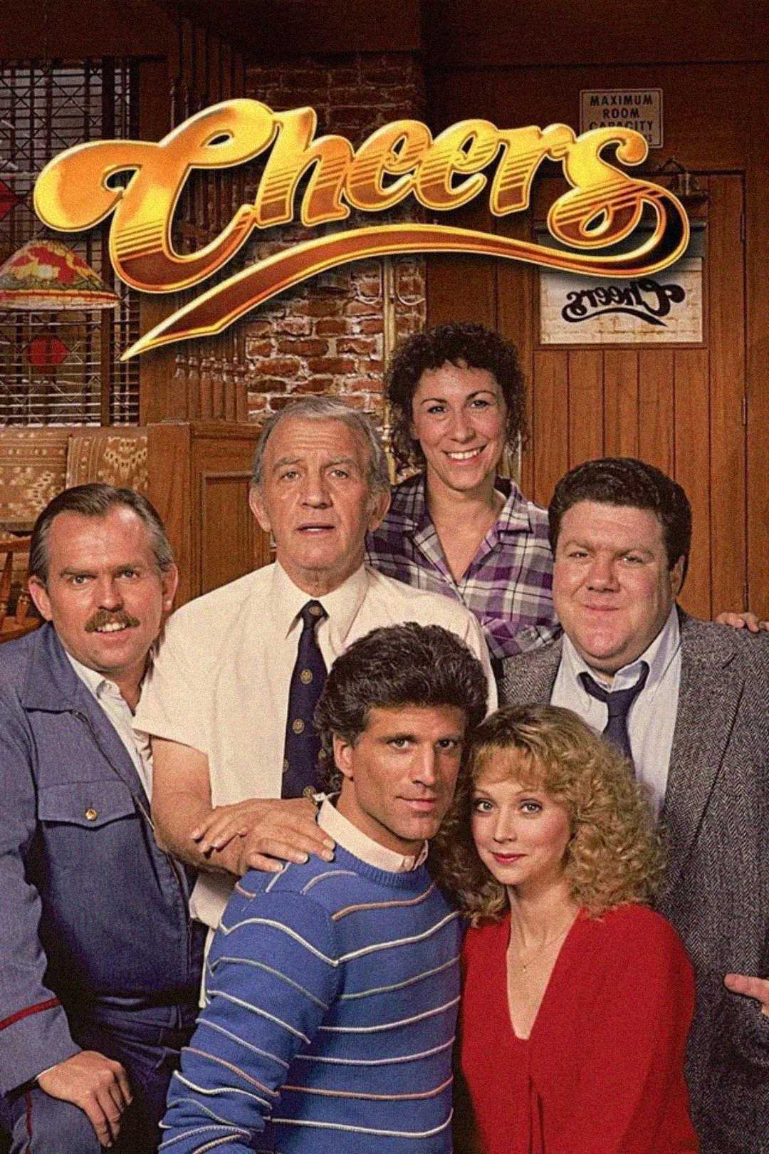 Six people pose together in a bar setting beneath the yellow "Cheers" logo; the group includes men and women, smiling and standing close to each other in front of wood-paneled walls and a stained glass lamp.