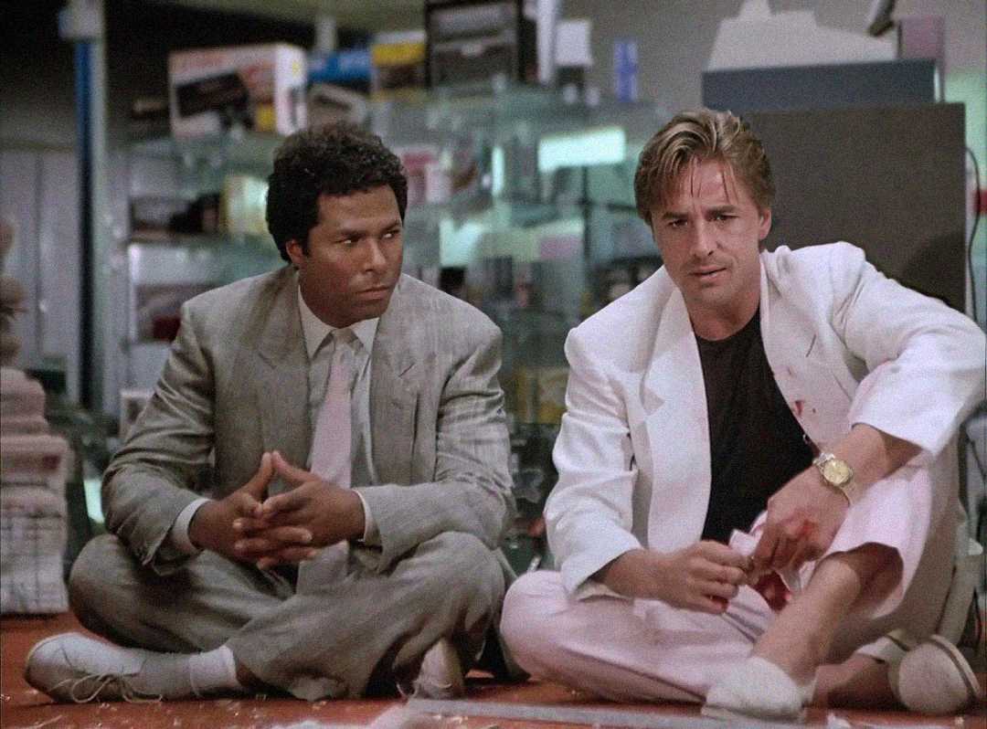 Two men in pastel suits sit on the floor of what appears to be an electronics store. One man wears a grey suit and tie, the other a white suit with a black shirt. Both appear serious and focused.