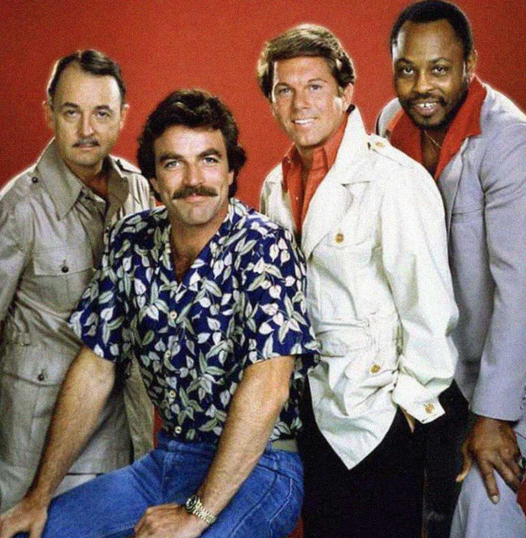 Four men pose together in front of a red background. One wears a floral shirt and sits on a stool, while the others stand around him in light-colored jackets. They all smile at the camera.