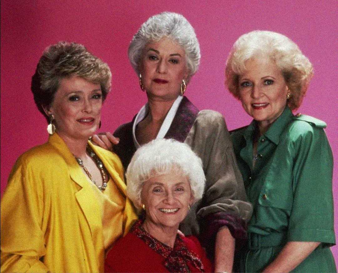 Four older women pose together against a pink background. Three stand in back wearing yellow, green, and gray jackets, while one sits in front wearing a red top. They all smile warmly at the camera.