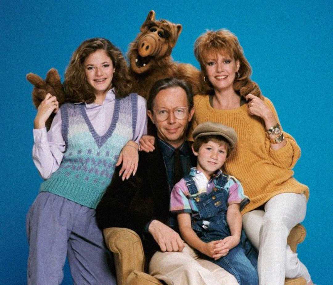 A family of four poses with a brown alien puppet against a blue background. The adults and children are smiling; the alien stands behind them, resting its hands on their shoulders.