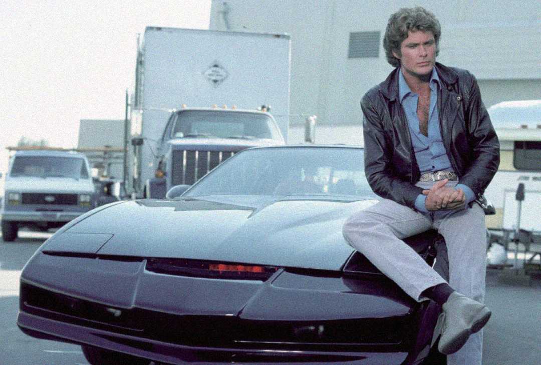 A man in a leather jacket and light pants sits on the hood of a sleek black car with a red light on the front, parked in a lot with trucks and trailers in the background.