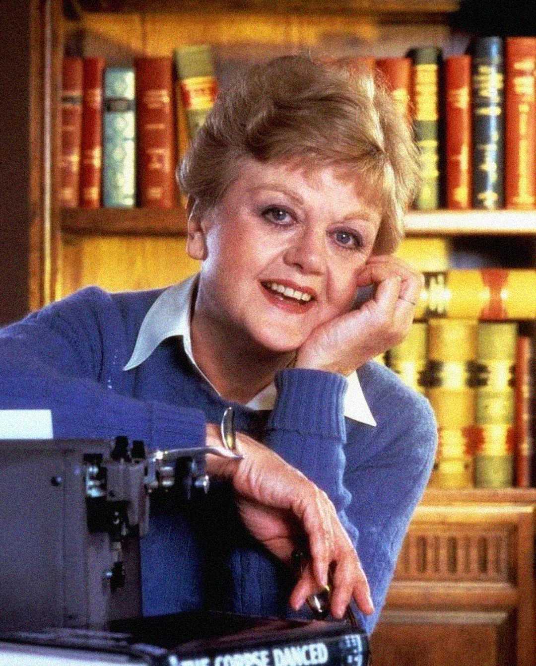 A smiling older woman with short blond hair sits by a typewriter, resting her head on her hand, with shelves of books in the background. She is wearing a blue sweater over a collared shirt.