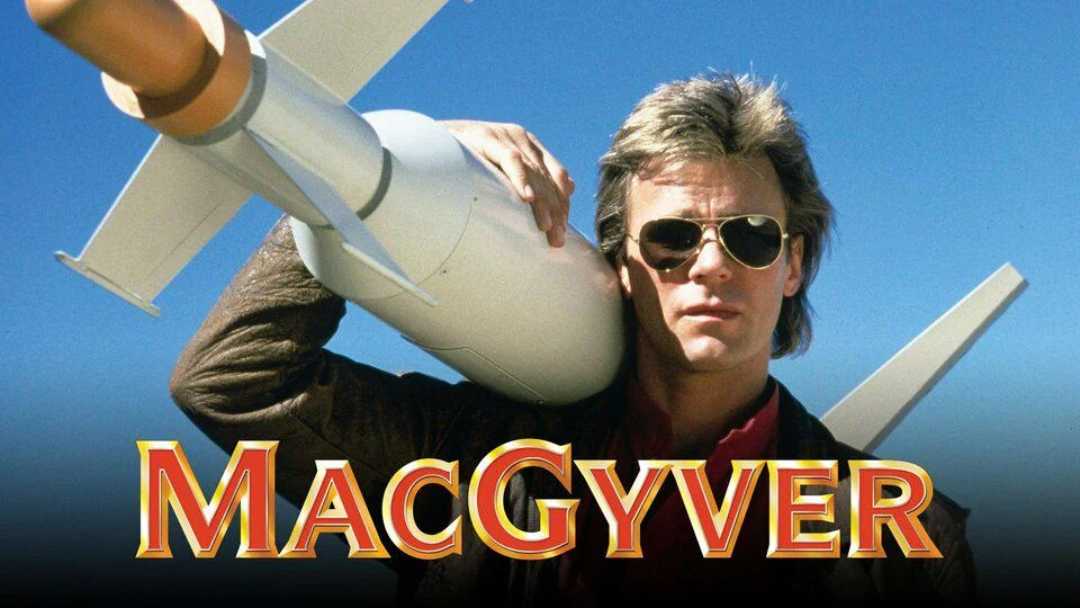 A man in sunglasses holds a large rocket on his shoulder against a blue sky. The word "MacGyver" appears in bold red and yellow letters at the bottom of the image.