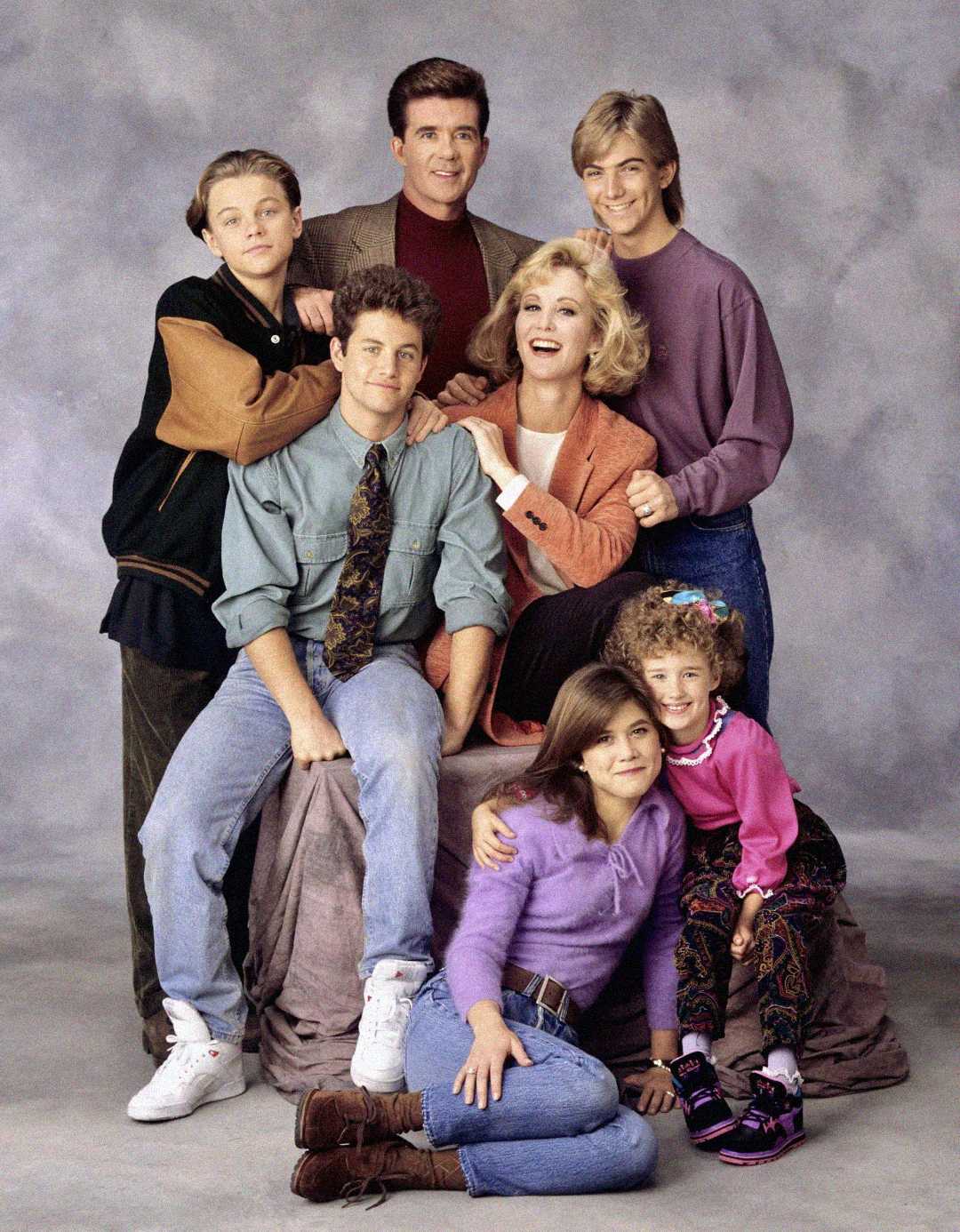 A group of seven people pose together, smiling, in front of a gray backdrop. Three adults stand and sit in the middle, surrounded by four children and teens in colorful 80s-90s style clothing.