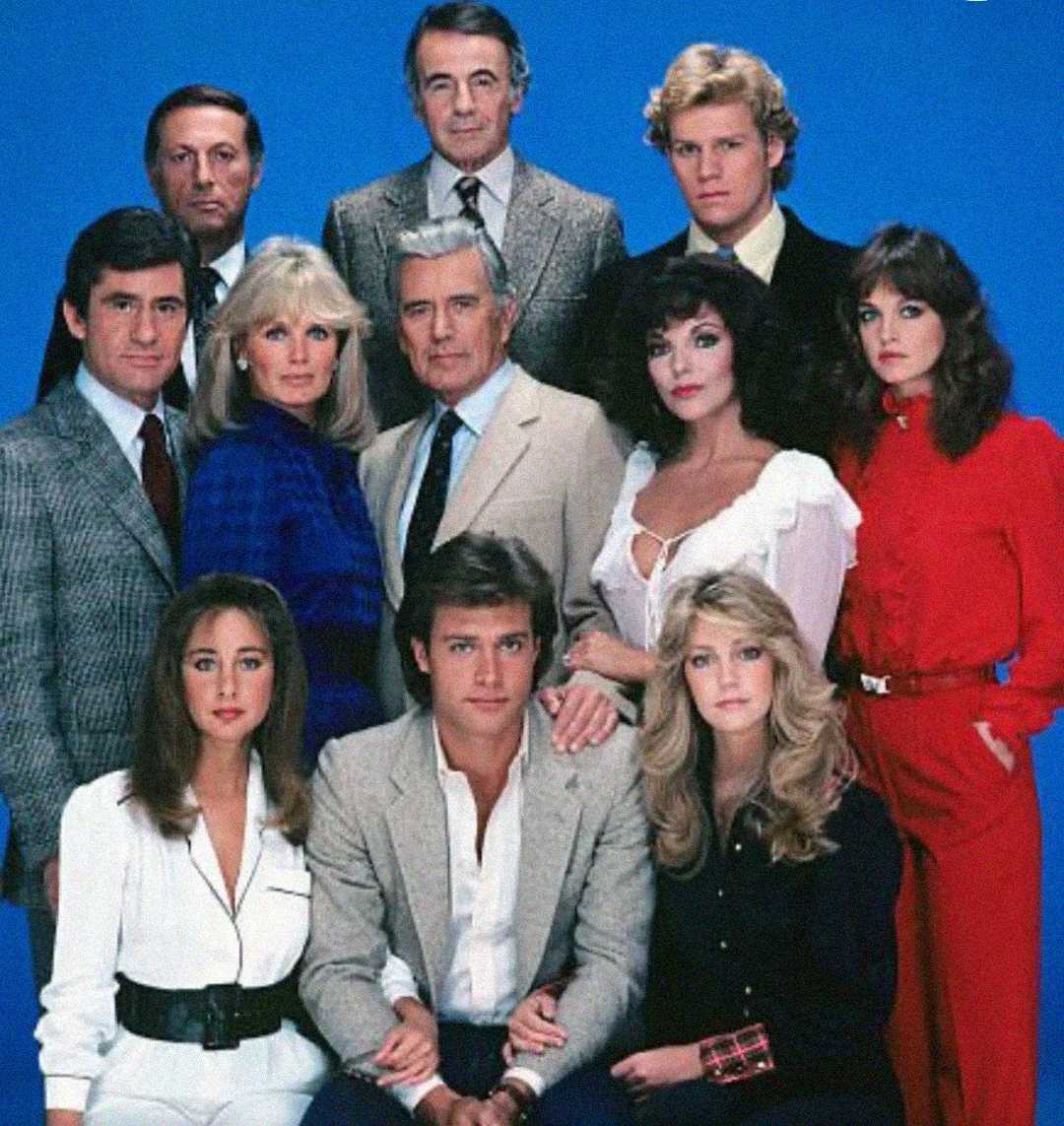 Twelve people, dressed in formal 1980s-style clothing, pose together against a solid blue background in three rows, with serious and neutral expressions.