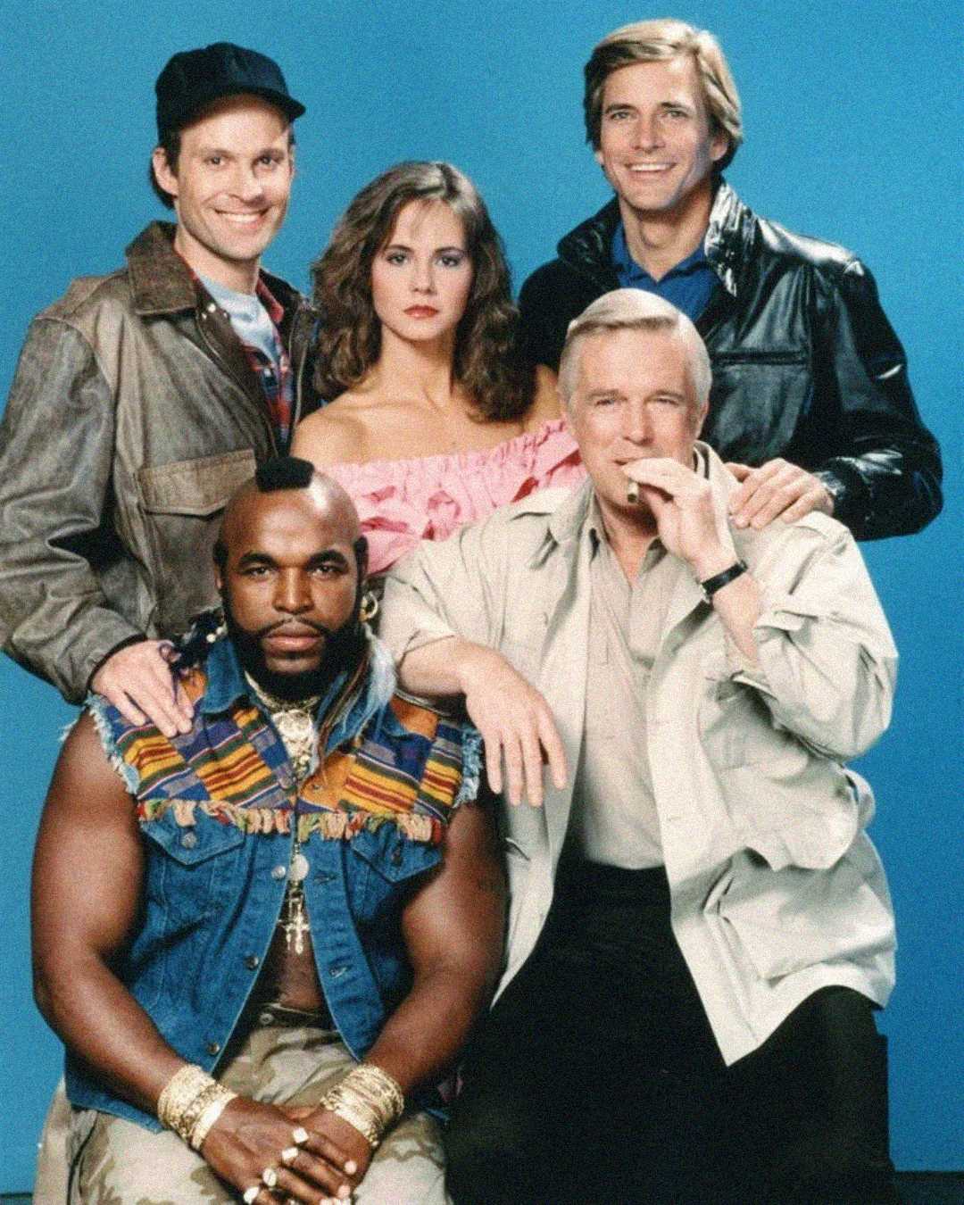 Five people pose together against a blue background; one man sits in front wearing gold chains and a colorful vest, while another man sits beside him smoking a cigar. Three people stand behind, smiling at the camera.