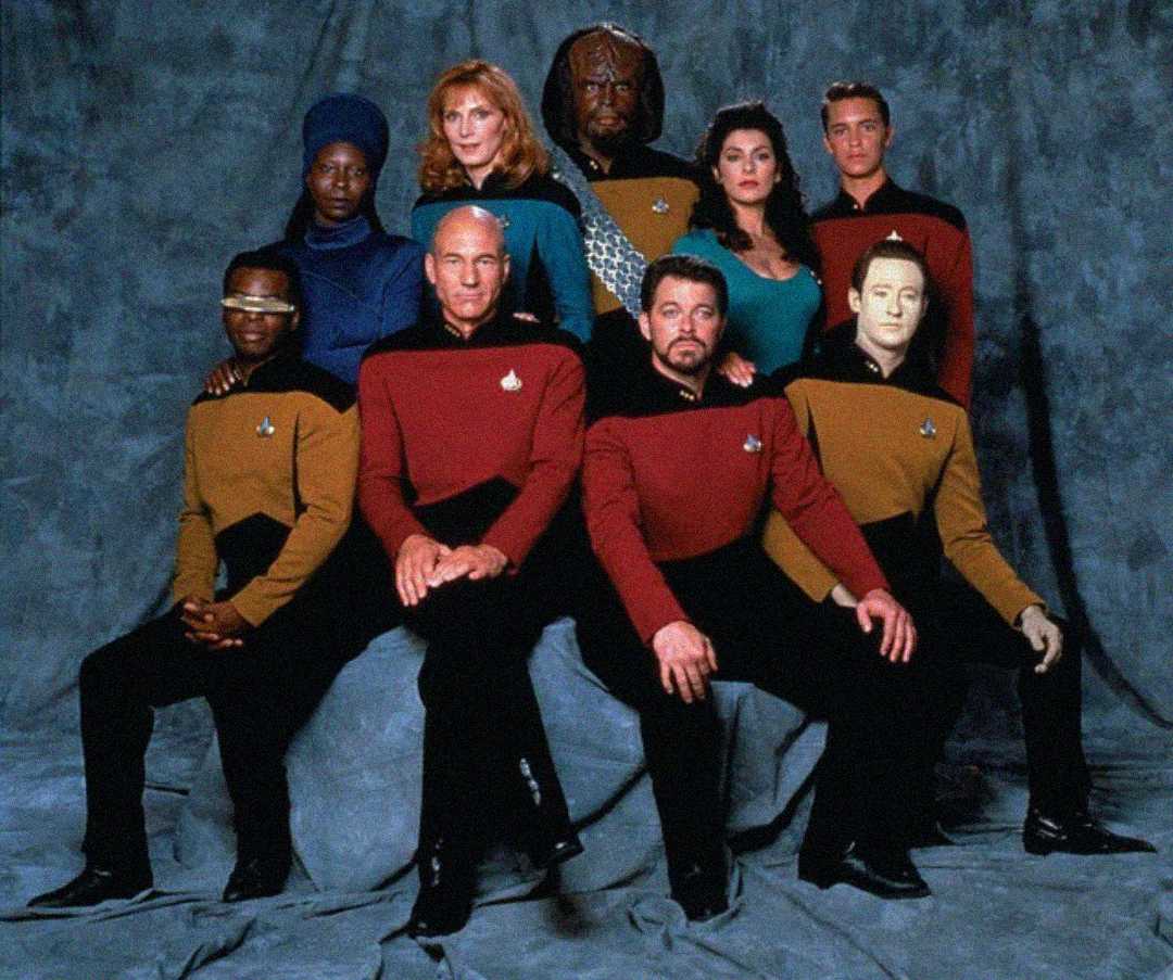 Nine people in Star Trek uniforms pose for a group photo against a gray backdrop, with some sitting and others standing; costumes are red, gold, and blue, and one has alien makeup and another has a metallic visor.