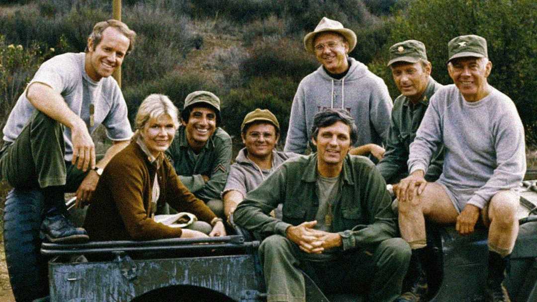 Eight people dressed in military-style clothing and casual outdoor wear sit and stand around a jeep, smiling at the camera with a background of trees and a hillside.