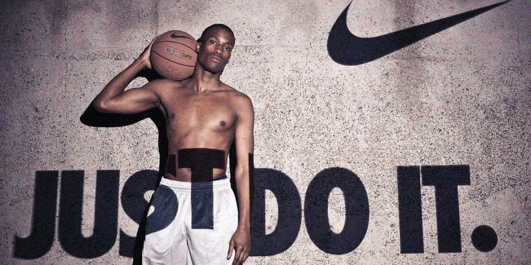 A shirtless man holding a basketball against his shoulder stands in front of a wall with the Nike logo and large text reading "JUST DO IT." He is wearing athletic shorts.