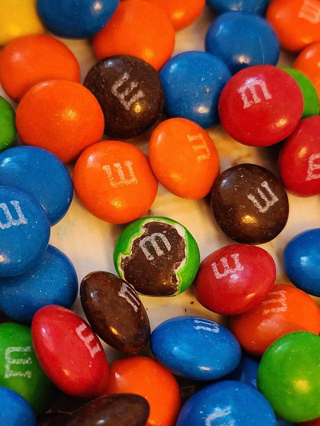 A pile of colorful M&M's candies, mostly red, blue, orange, brown, and green. One green candy in the center has a bite taken out, exposing the chocolate inside.