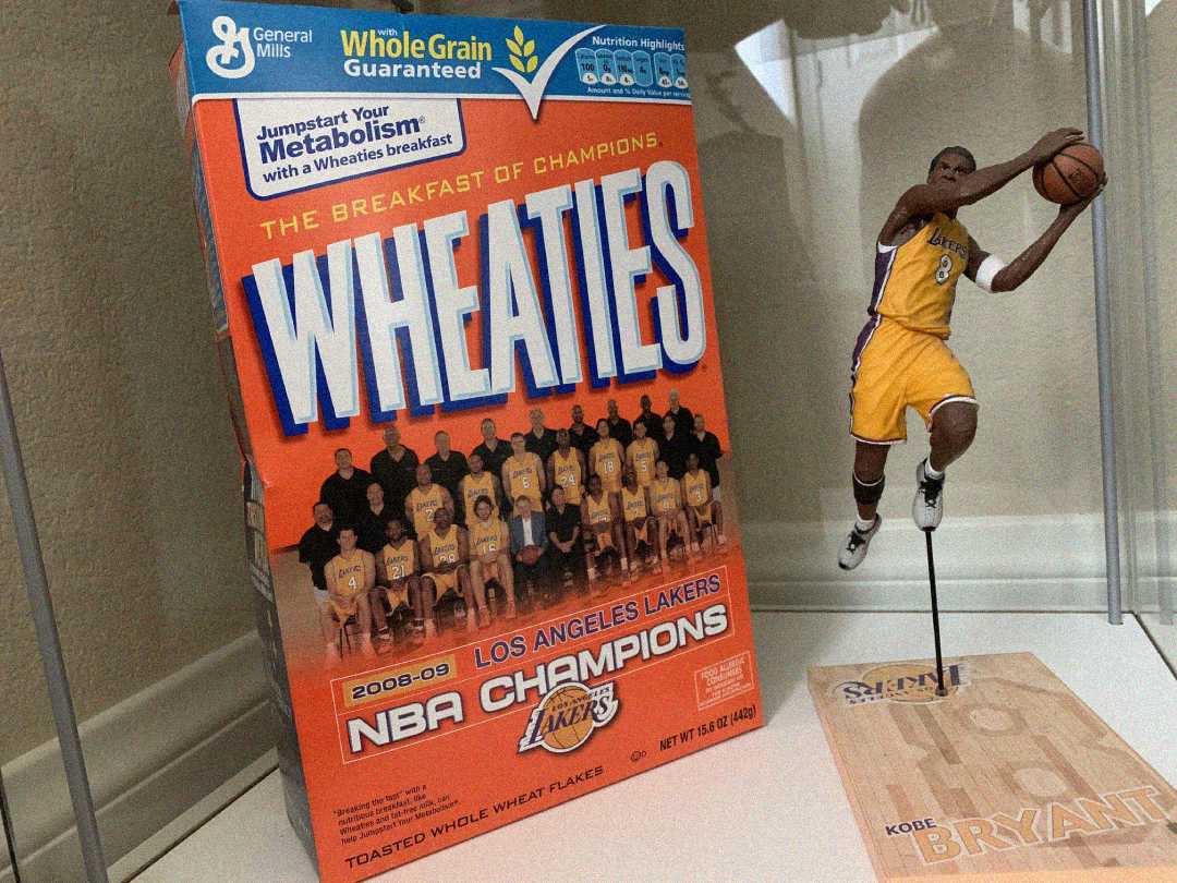 A Wheaties cereal box featuring the 2008–09 NBA Champion Los Angeles Lakers team photo, with a small figurine of Kobe Bryant in mid-jump shot displayed beside it.