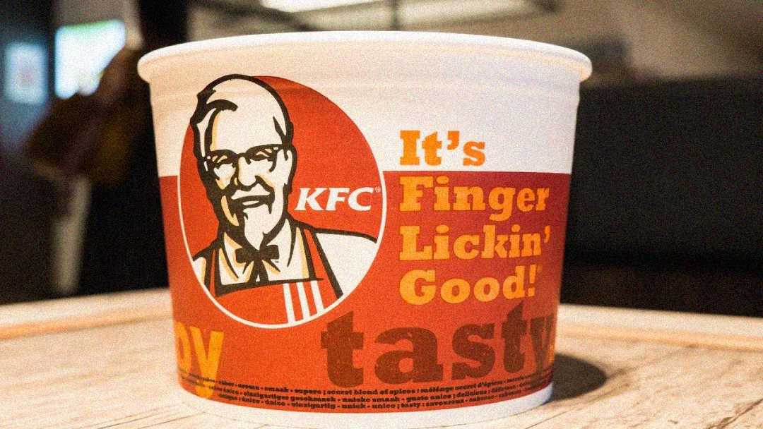 A close-up of a KFC bucket featuring the Colonel Sanders logo and the slogan "It’s Finger Lickin’ Good!" in bold orange and yellow text, sitting on a light-colored table.