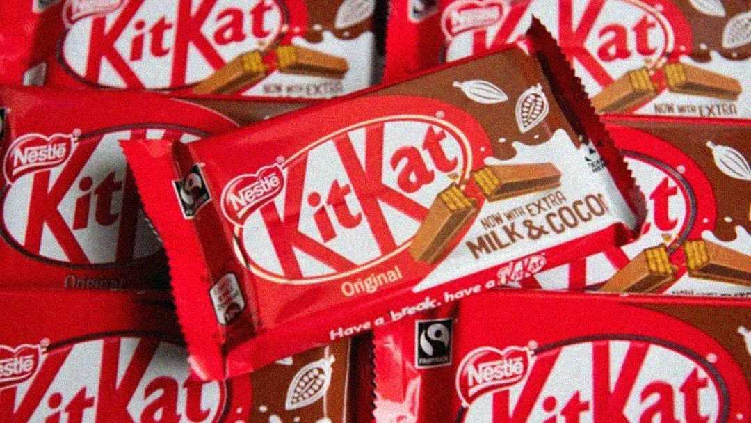 Several red-wrapped Kit Kat chocolate bars are stacked together, with the largest one in the center showing “Nestlé Kit Kat Original” and “Now with Extra Milk & Cocoa” text on the packaging.