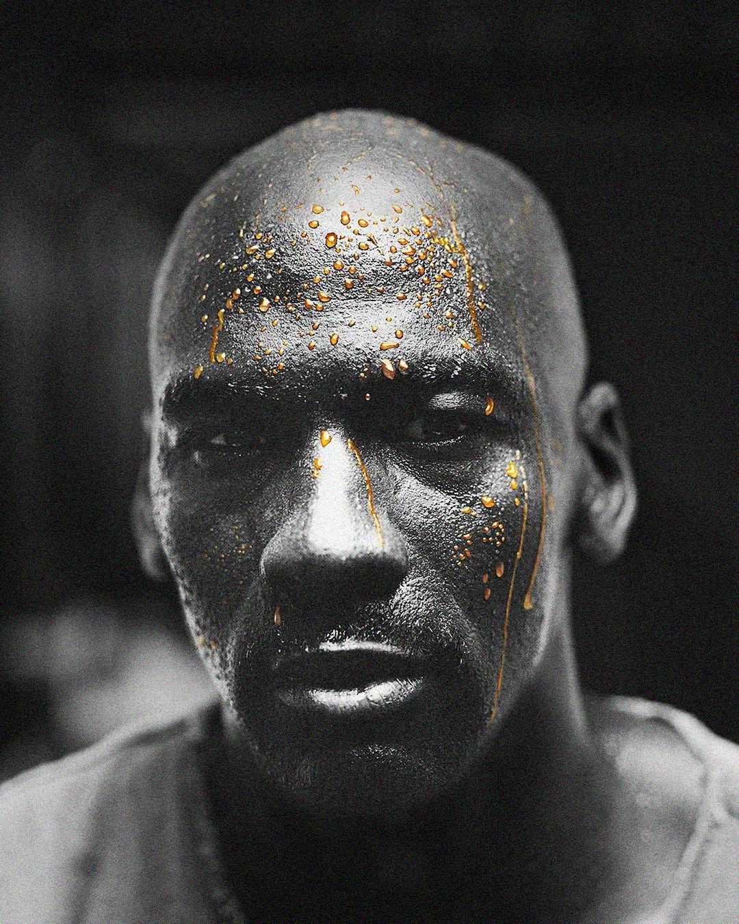 A close-up, black and white portrait of a man with a shaved head. Gold liquid drips across his face and scalp, creating a striking contrast against the dark background and his serious expression.