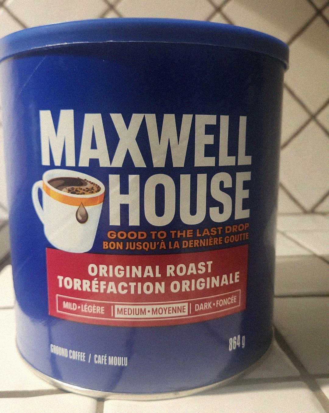A blue container of Maxwell House Original Roast ground coffee sits on a tiled countertop. The label includes both English and French text and shows a steaming coffee cup. The container holds 864 grams of coffee.