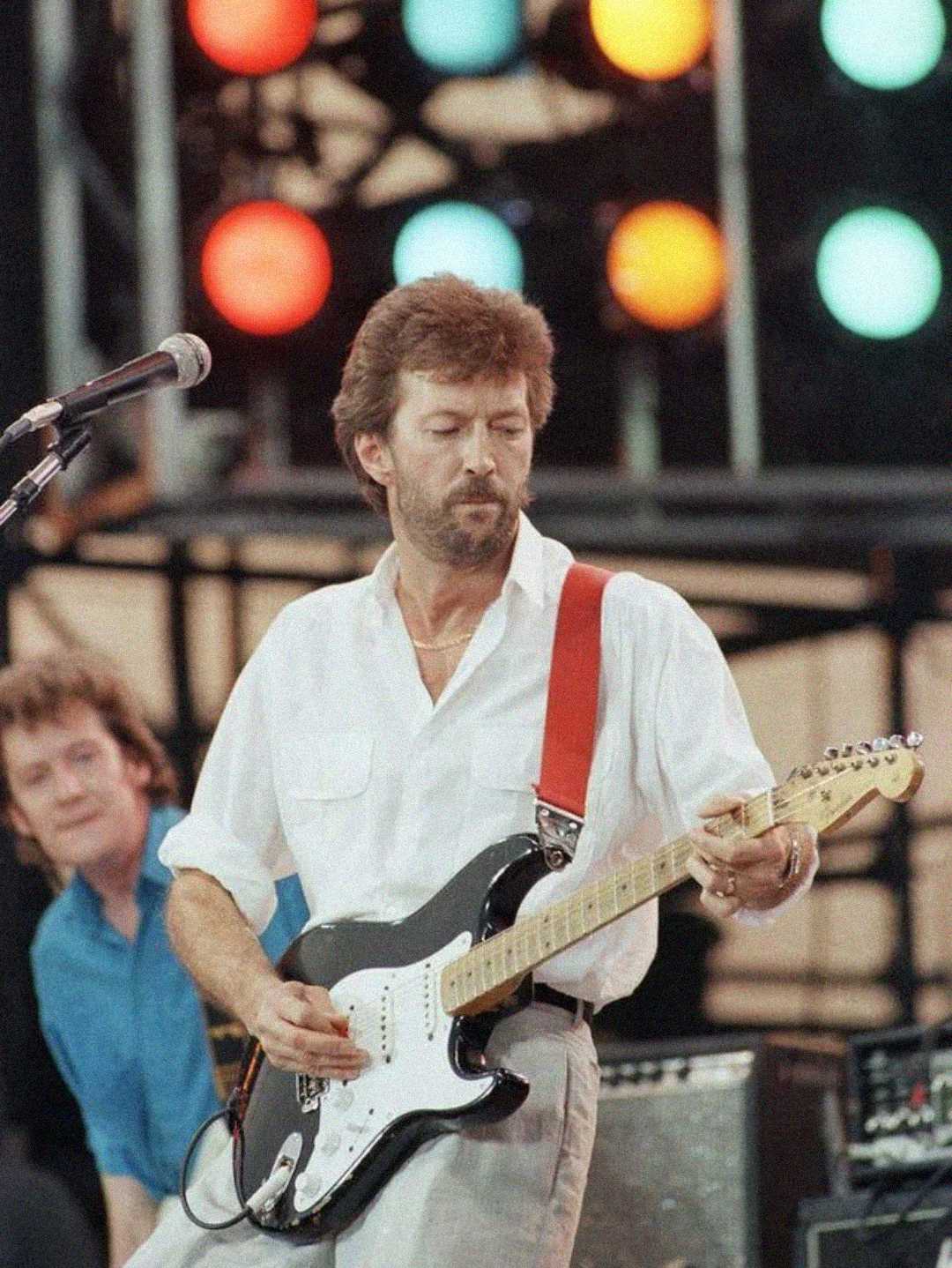 A man in a white shirt plays an electric guitar on stage, with colorful stage lights in the background and another musician visible behind him.