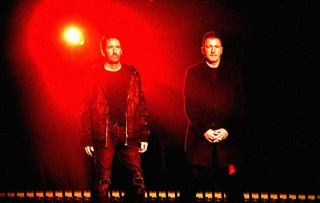 Two men stand side by side under dramatic red lighting, with a bright light source behind them. Both are dressed in dark clothing, creating strong silhouettes against the glowing background.