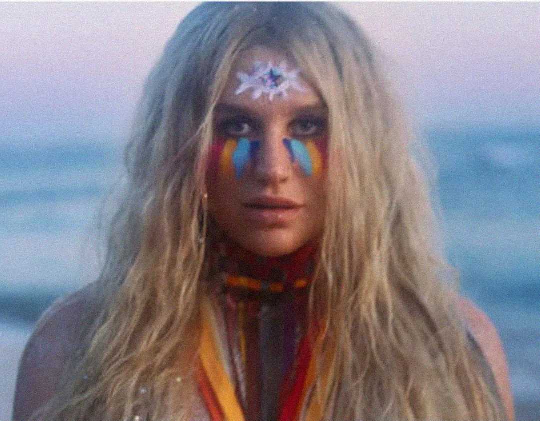 A person with long blonde hair stands on a beach at sunset, wearing colorful face paint with blue, red, and yellow stripes under their eyes and a white star design on their forehead. They have bright ribbons around their neck.