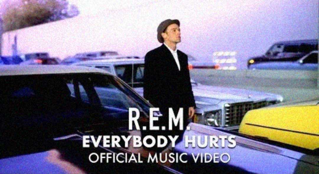 A man in a hat stands between cars in a parking lot at dusk. Text reads: "R.E.M. Everybody Hurts Official Music Video.
