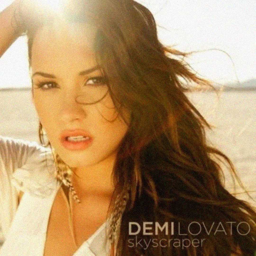 A woman with long, wavy hair poses outdoors in sunlight, gazing at the camera. Text at the bottom reads "DEMI LOVATO skyscraper." The background appears to be a bright, sandy landscape.