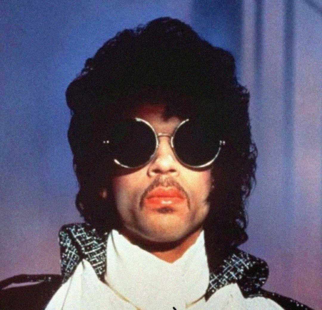 A person with curly dark hair, wearing large round sunglasses, a high-collared jacket, and a serious expression, stands against a blurred background.
