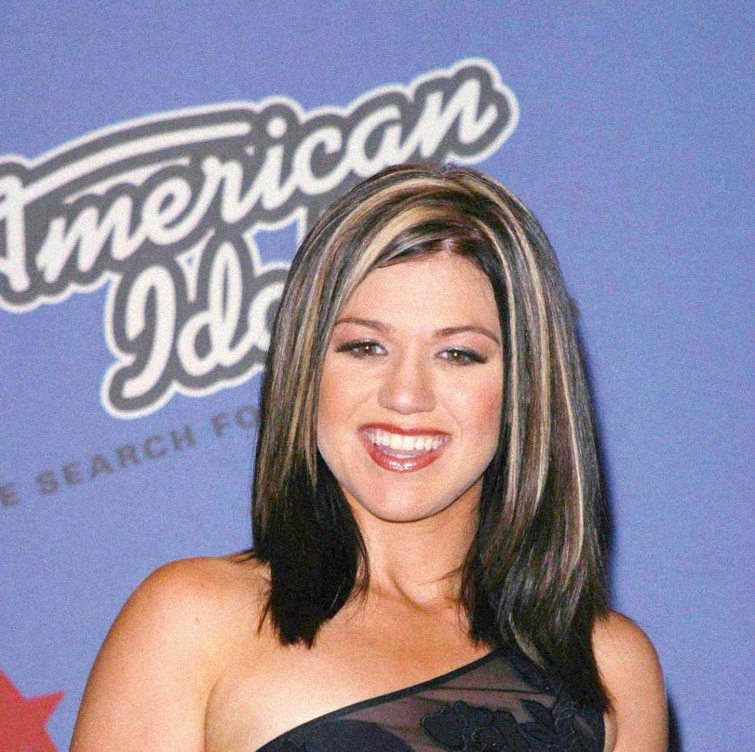 A woman with shoulder-length dark hair featuring blonde highlights smiles in front of a blue American Idol backdrop. She is wearing a one-shoulder black dress.