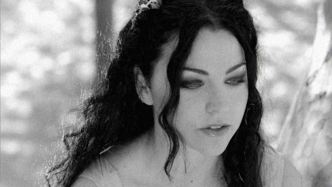 A woman with long, dark wavy hair and dramatic eye makeup looks downward in a black-and-white photo. Light filters through trees in the background, creating a soft, ethereal atmosphere.