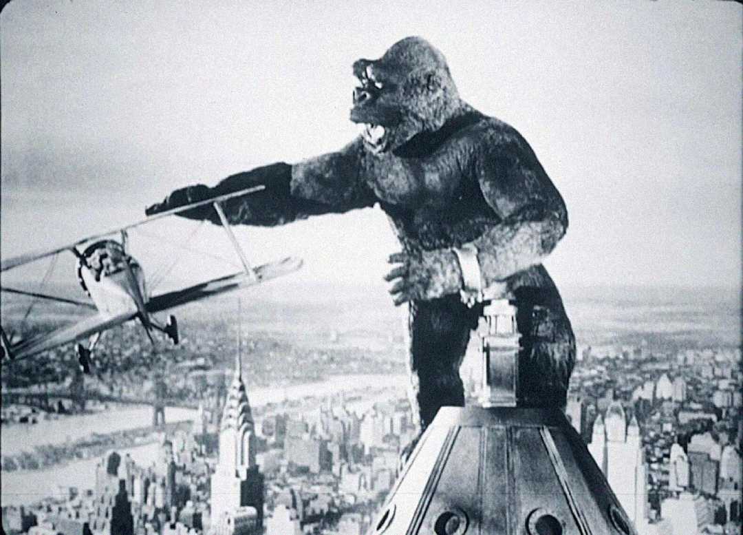 A giant gorilla stands atop a tall skyscraper, reaching out to swat a biplane flying nearby, with a city skyline visible in the background.