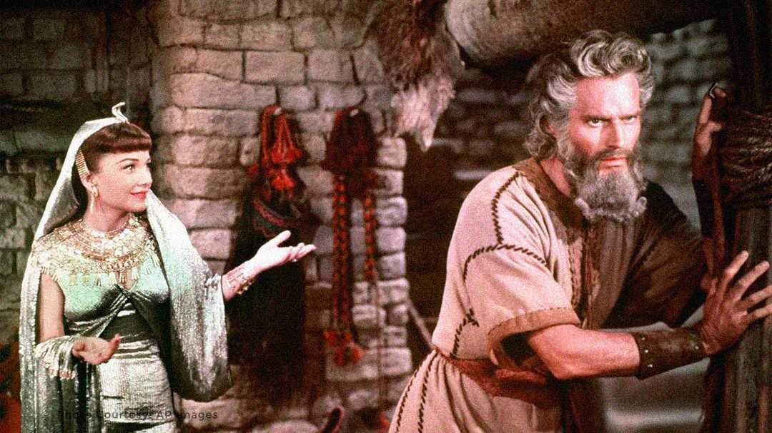 A woman in ancient Egyptian attire gestures toward a bearded man in rustic robes, who looks serious and holds onto a wooden post in a stone-walled room decorated with draped fabrics and objects.