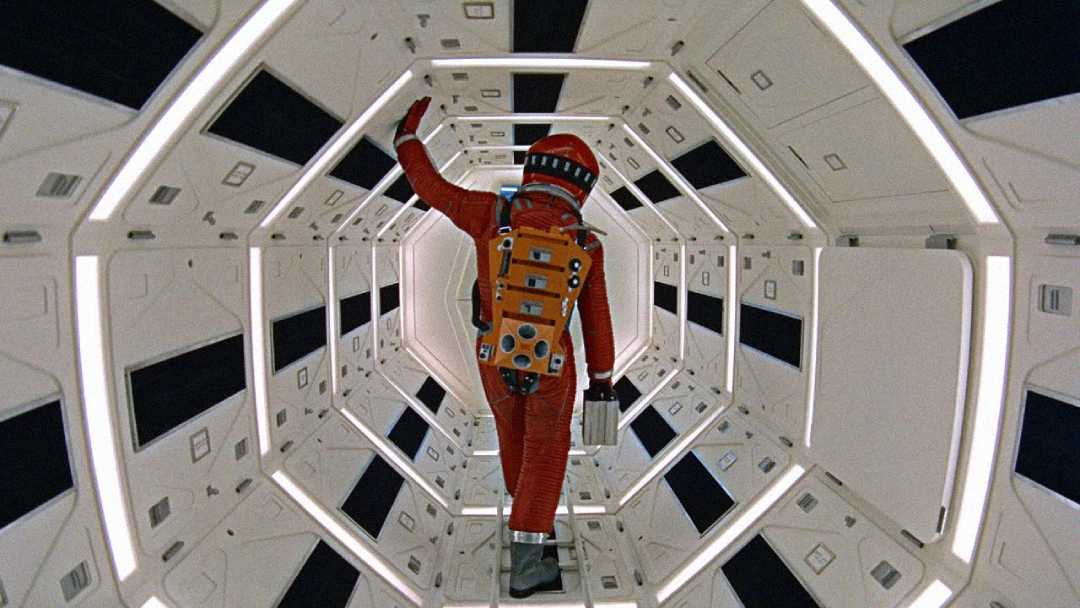 An astronaut in a red spacesuit walks through a brightly lit, futuristic, hexagonal corridor with white walls and black panels, holding onto a handrail above.