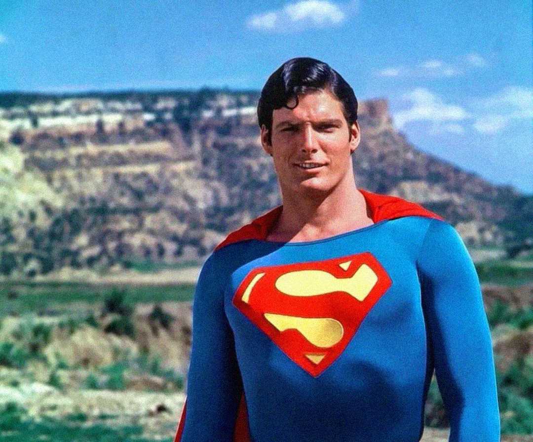 A man in a bright blue Superman costume with a red cape and yellow "S" emblem stands outdoors in front of a rocky landscape under a blue sky with clouds.