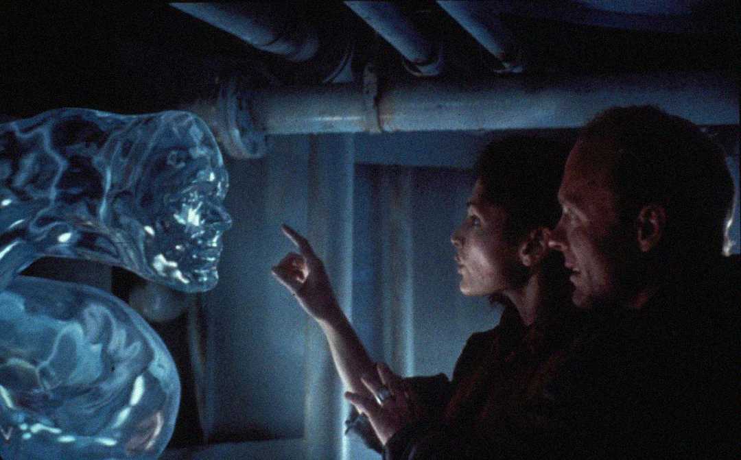 A woman reaches out to touch a translucent, water-like humanoid figure, as a man stands beside her in a dimly lit, industrial setting with pipes overhead.