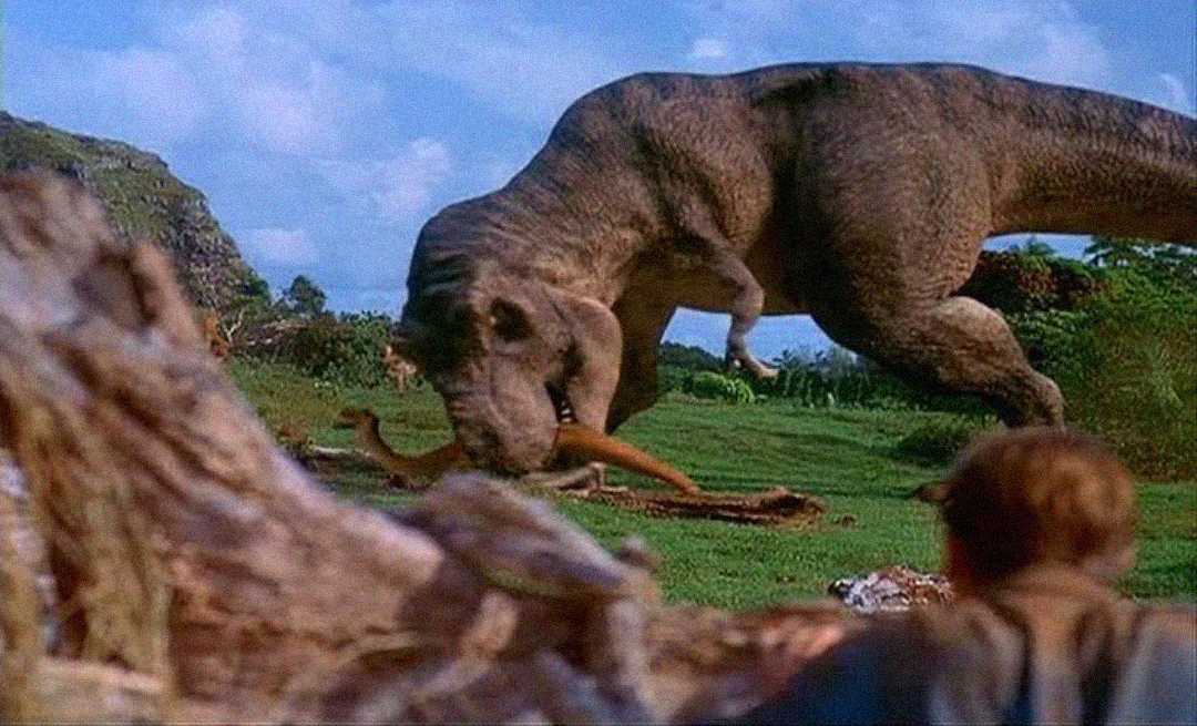 A large Tyrannosaurus rex bends down to eat another dinosaur in a lush, green landscape, while a person crouches in the foreground, partially hidden behind a log.
