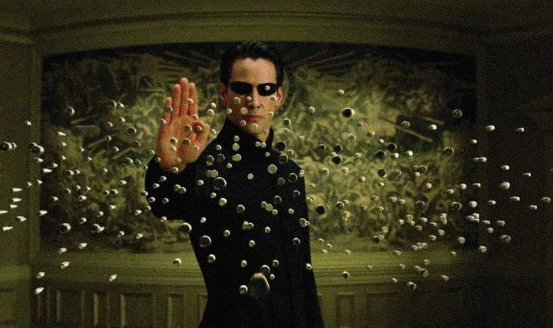 A person in a black coat and sunglasses stands with one hand raised, stopping a swarm of bullets in mid-air in a dramatic, dimly lit room with a patterned wall behind them.