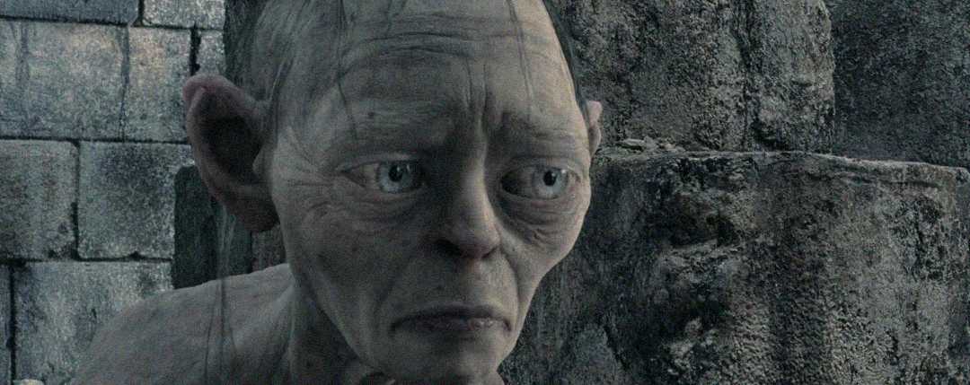A CGI character with pale skin, big blue eyes, and sparse hair looks sad and worried, sitting against a backdrop of gray stone walls.