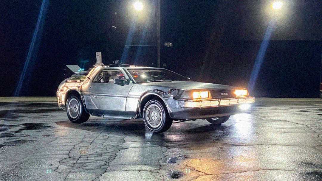 A silver DeLorean sports car with headlights on is parked on a wet, cracked pavement at night, illuminated by bright overhead lights. The car's gullwing door is open.