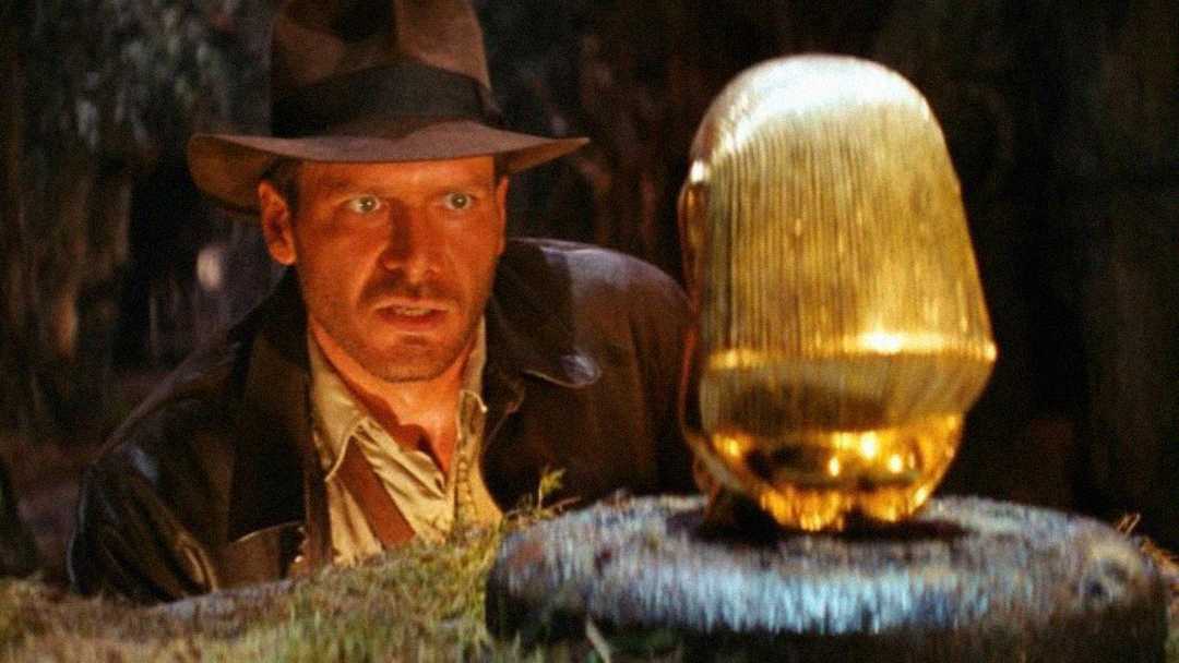 A man in a fedora and leather jacket intently stares at a shiny golden idol on a stone pedestal in a dim, jungle-like setting.