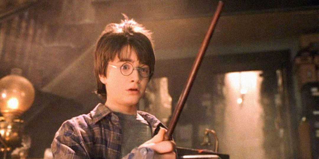A young boy with round glasses and messy brown hair holds a wand while wearing a plaid shirt. He looks surprised, standing indoors with soft light shining behind him and shelves in the background.