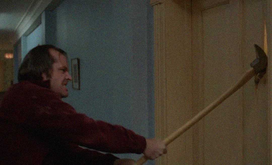 A man with a furious expression swings an axe, breaking through a wooden door in a dimly lit hallway.