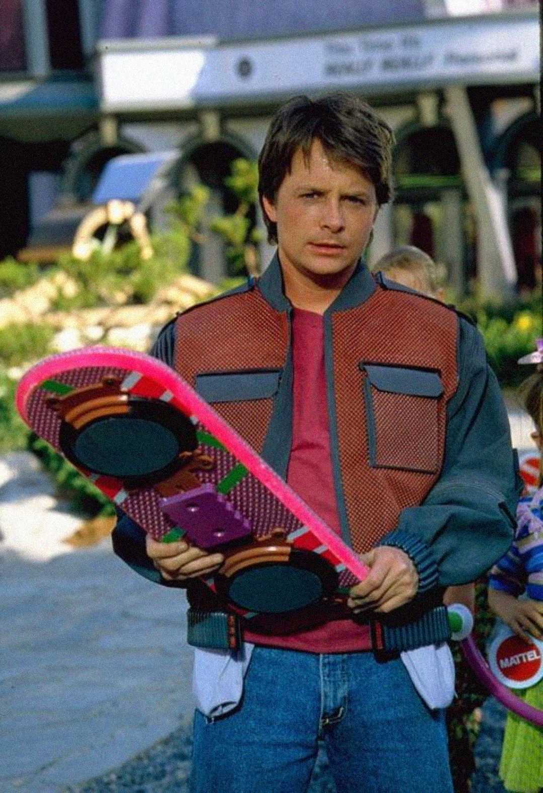 A young man in a red and gray futuristic jacket holds a colorful hoverboard with pink edges. He stands outdoors, looking directly at the camera, with blurred buildings in the background.