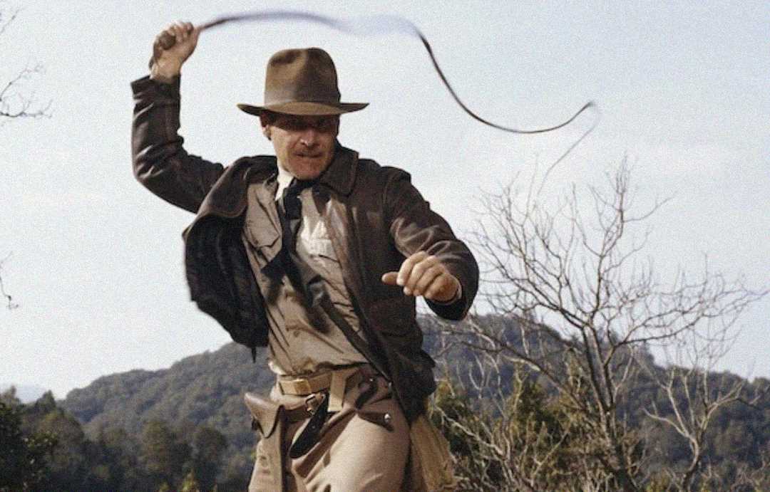 A man wearing a brown fedora, leather jacket, and khaki clothing swings a whip outdoors, with trees and hills in the background.
