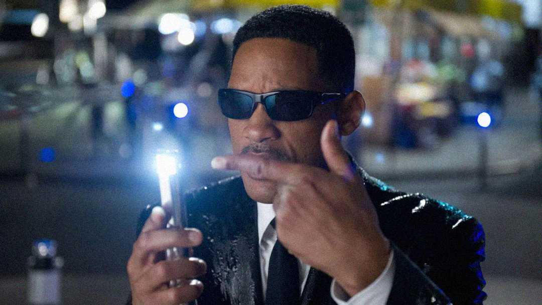 A man in a black suit and dark sunglasses holds up a small, pen-like device with a bright light, pointing at it. The background is blurred, suggesting a nighttime or urban setting.