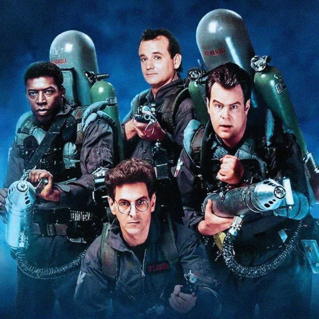 Four men in gray jumpsuits with proton packs pose together against a blue background, holding ghost-catching equipment. Their expressions are serious and alert. This is a promotional image from the movie "Ghostbusters.