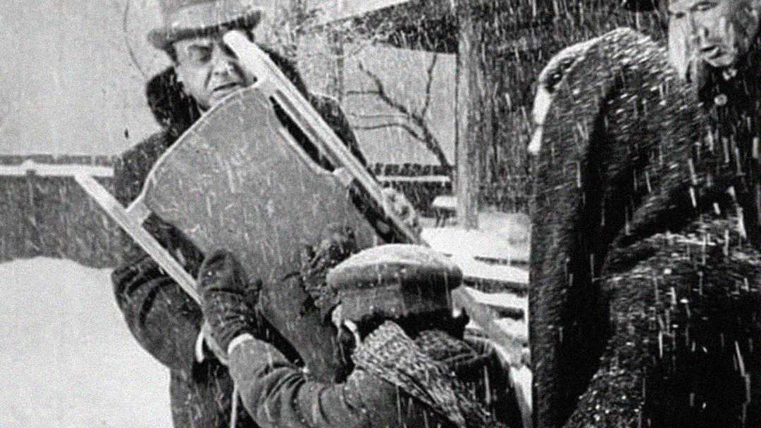 A black-and-white photo shows people outdoors in heavy snowfall. One person holds a sled upright while a child, bundled in winter clothes, interacts with it. Other adults, also dressed warmly, stand nearby. Snow covers the ground and falls thickly.
