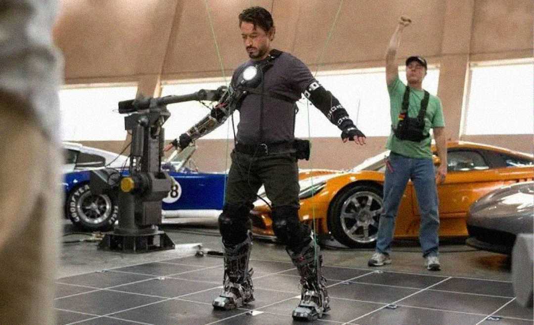 A man wearing a motion capture suit with sensors stands in a garage next to sports cars, while another person in the background raises a fist in the air.