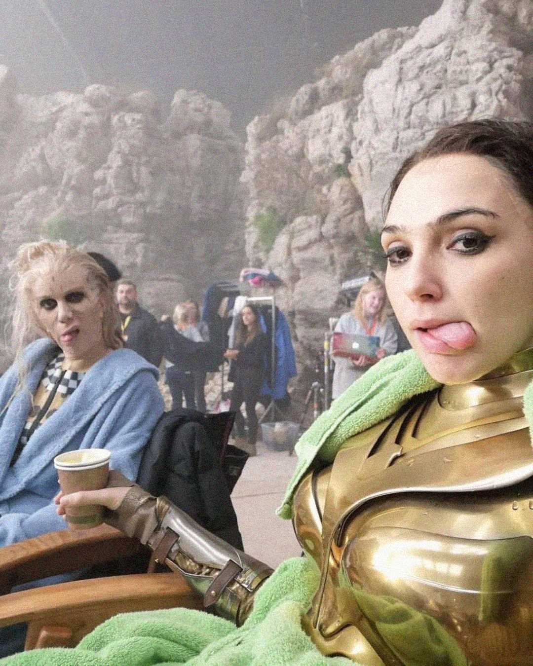 Two women in costume relax on set; one wears gold armor and a green blanket, sticking out her tongue, while the other has wild hair, pale makeup, and holds a cup. Rocky set background with crew members visible behind them.